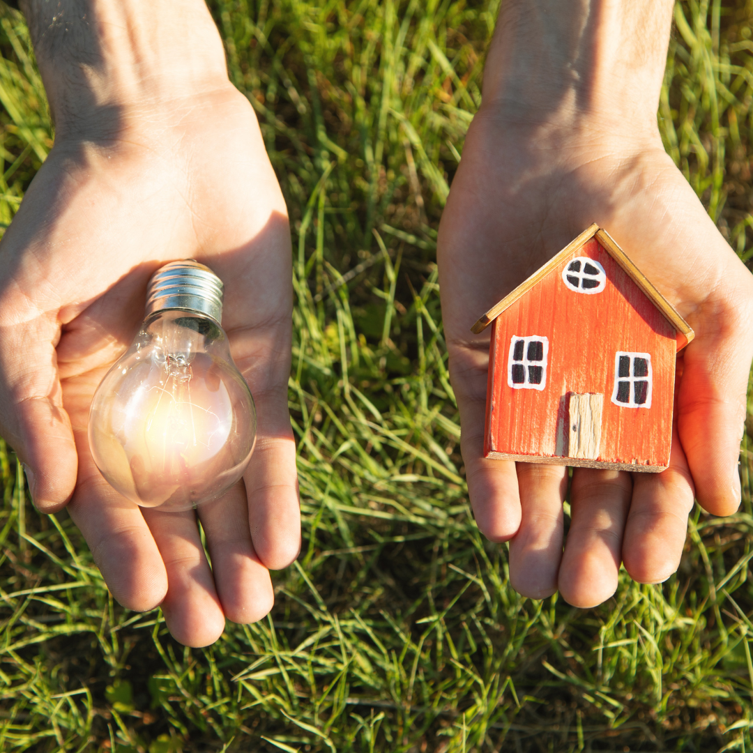 Two hands holding a lightbulb and a small model house, symbolizing energy and housing solutions, with a grassy background.